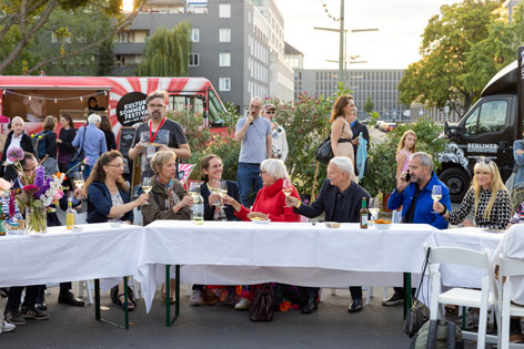 Kulturstaatsministerin Claudia Roth und Landwirtschaftsminister Cem Özdemir (links hinter Blumenstrauß) tafeln in der Sigismundstraße mit Anrainer*innen und Gäst*innen © Stefanie Heider Menschen prosten sich an einer Tafel im Freien gutgelaunt zu