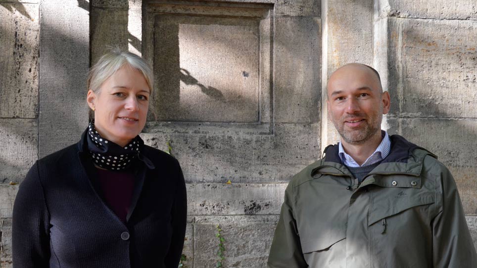 Nina Schallenberg und Daniel Naumann, Nachhaltigkeitsbeauftragte der SPK Eine Frau und ein Mann stehend lächelnd vor einer grauen Mauer
