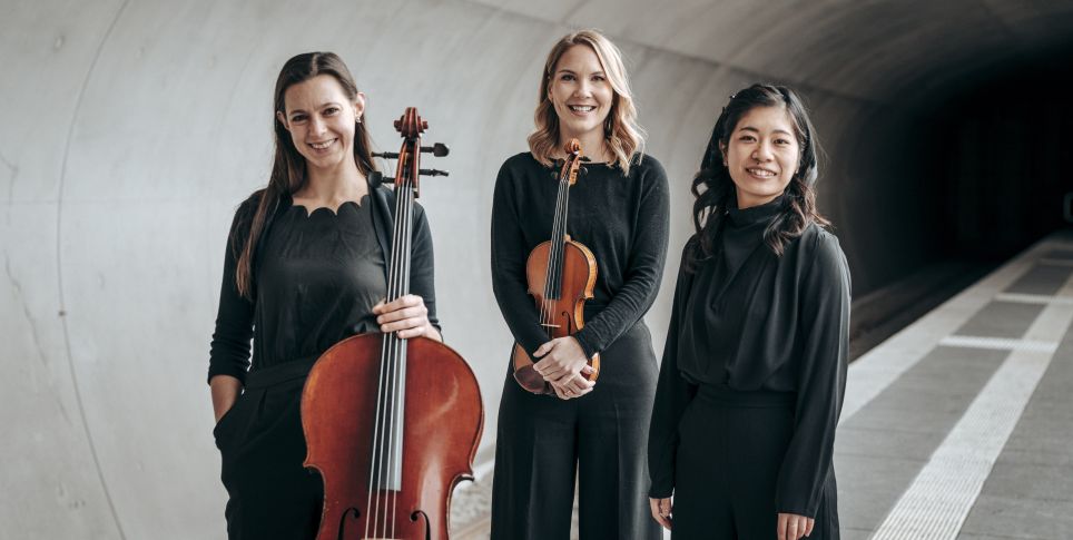 Die drei Musikerinnen von Trio Hannari stehen mit ihren Instrumenten und schwarzer Kleidung vor einer Betonwand