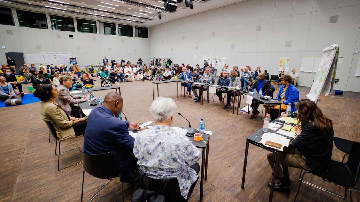 Workshop „Gemeinsam Arbeiten“ Mehrere Personen sitzen im Halbkreis an Tischen, im Hintergrund am Boden sitzende Personen, die der Diskussion lauschen