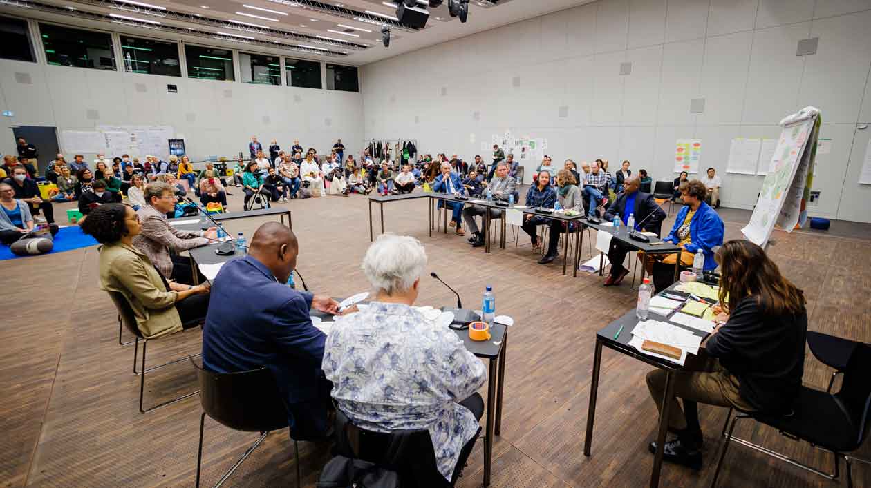 Several people are sitting in a semicircle at tables, in the background people are sitting on the floor listening to the discussion