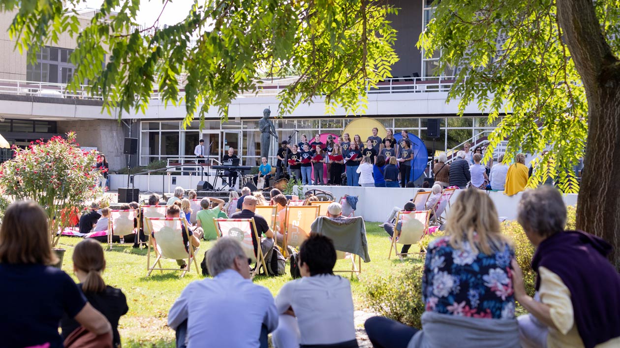 Menschen sitzen auf einer Grünfläche vor einem modernen Gebäude und lauschen einem Kinderchor