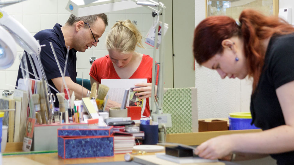 Auszubildende in der Buchbinderei der Staatsbibliothek Zwei Auszubildende und ihr Ausbilder arbeiten in der Buchbinderei