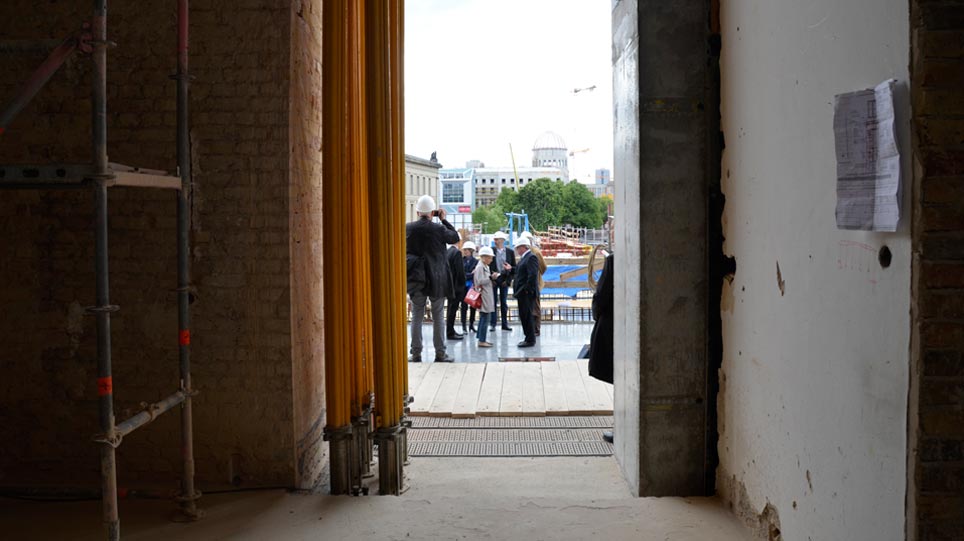 Blick von einer Baustelle durch eine Maueröffnung nach draußen, wo mehrere Menschen mit weißen Bauhelmen stehen (öffnet Vergrößerung des Bildes)