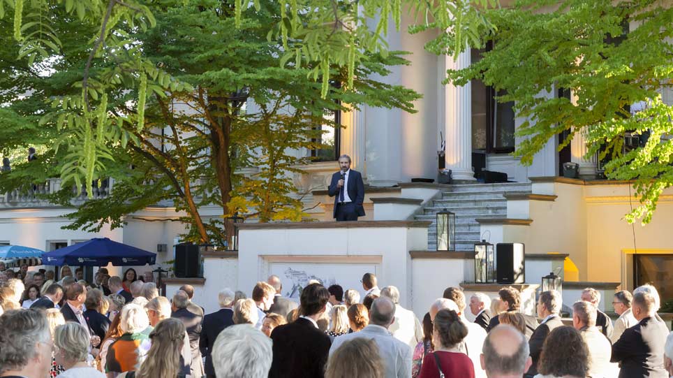 Ein Mann steht auf der Treppe einer Villa im Freien und spricht in ein Mikrofon zu einem Publikum