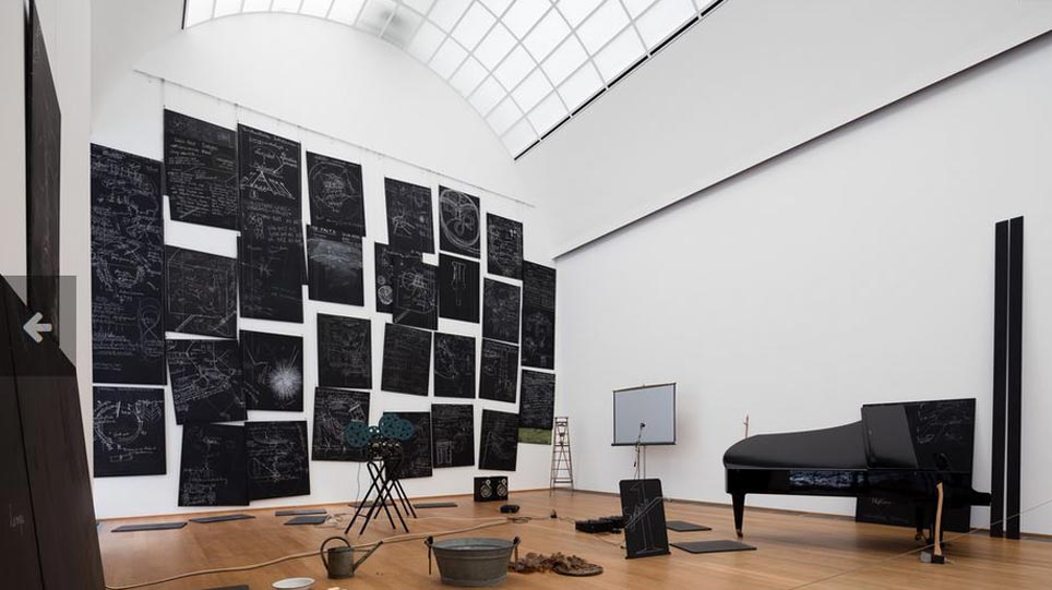 Künstlerische Installation mit Kreidezeichnungen an schwarzen Tafeln, einem schwarzen Flügel, einem Filmprojektor, einer Leiter und einer Zinkwanne in einem hohen Raum (öffnet Vergrößerung des Bildes)
