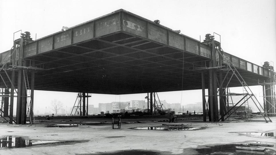 Historical photograph of the building of the Neue Nationalgalerie showing the steel roof being raised with hydraulic jacks (opens enlarged image)