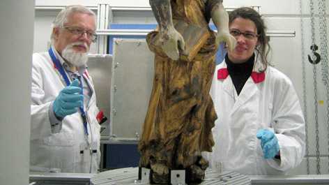 Zwei Personen in Laborkleidung betrachten eine Holzskulptur, die in einem Gerät untersucht wird
