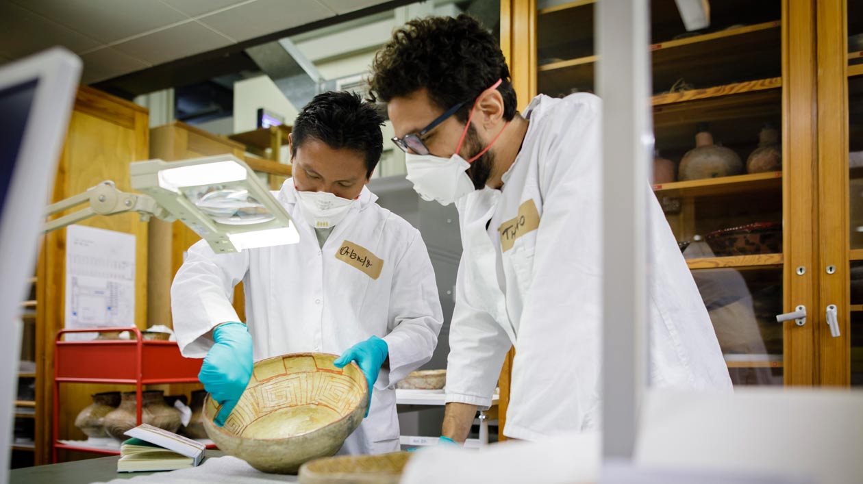 Untersuchen eine Baniwa-Keramik aus der Sammlung des Ethnologischen Museums: Thiago da Costa Oliveira (re.) und Orlando Fontes, ein Forscher und Vertreter von der indigenen Gruppe der Baniwa beim Workshop „Geteiltes Wissen“. Zwei Männer in Schutzausrüstung untersuchen eine Keramikschale