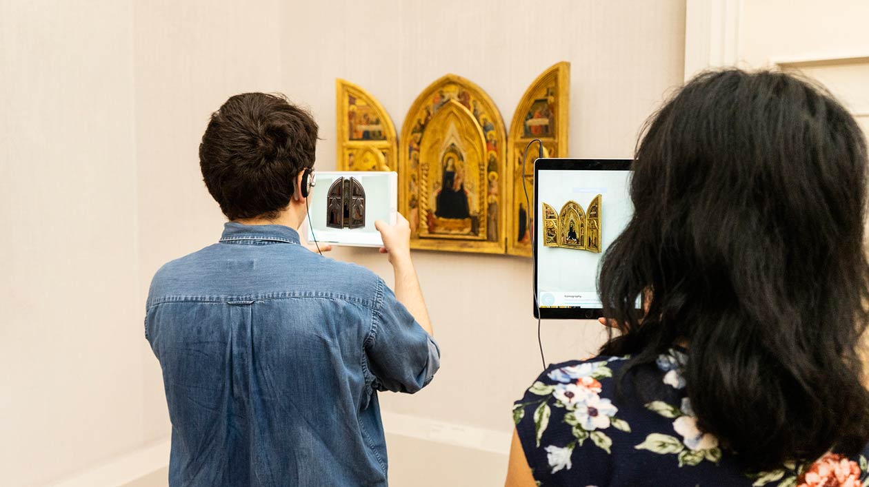 Zwei Personen halten Tablets vor ein Triptychon in einem Ausstellungssaal