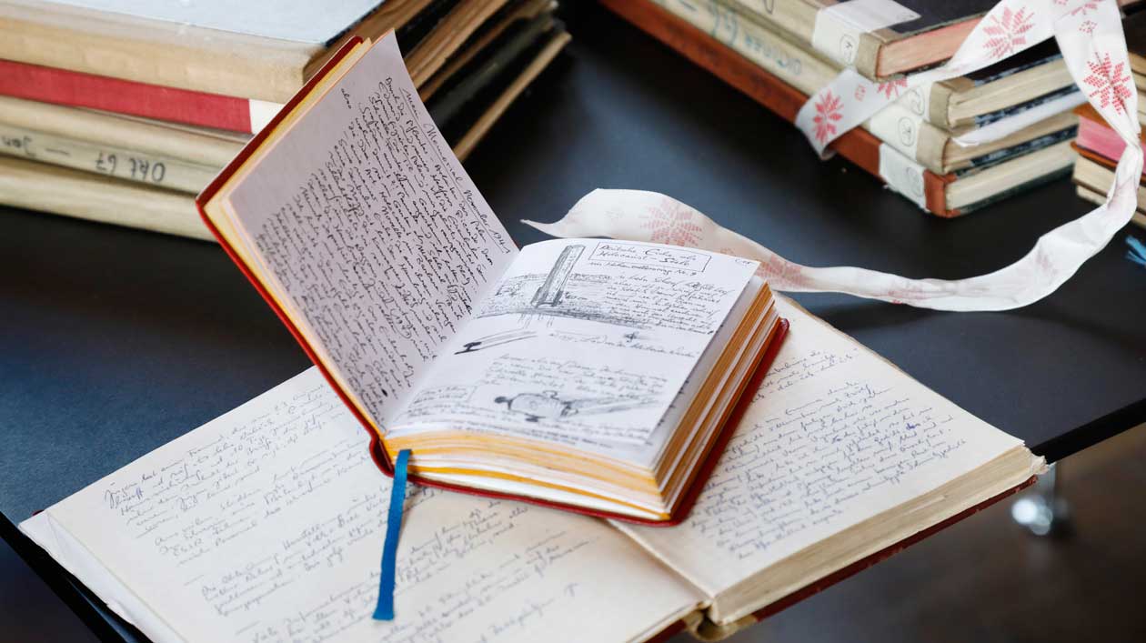 Ein Tagebuch liegt aufgeschlagen auf einem anderen aufgeschlagenen Tagebuch, im Hintergrund Bücherstapel 