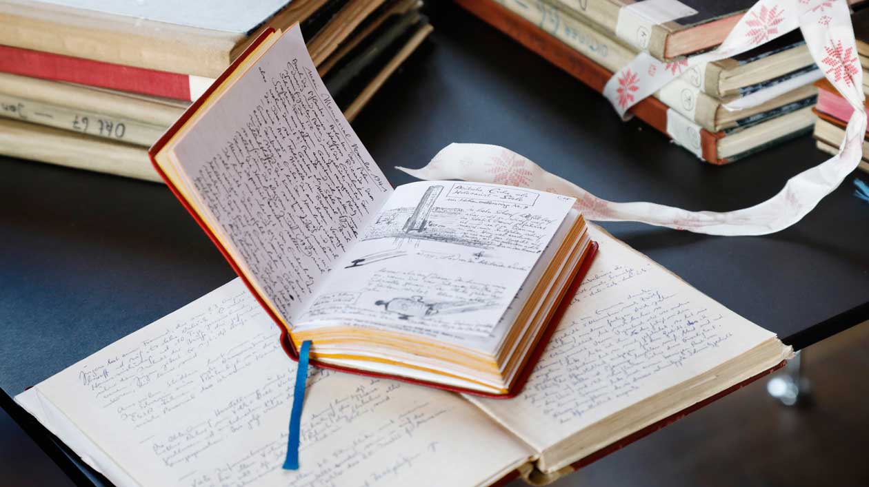 A diary lies open on top of another open diary, in the background stacks of books 