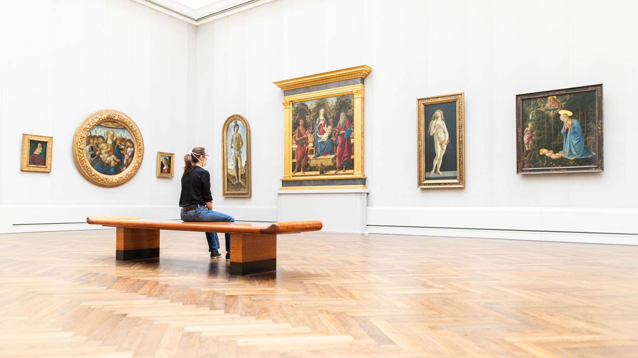 Eine Frau mit Mundschutz sitzt in einem Museum auf einer Holzbank und betrachtet mittelalterliche Gemälde