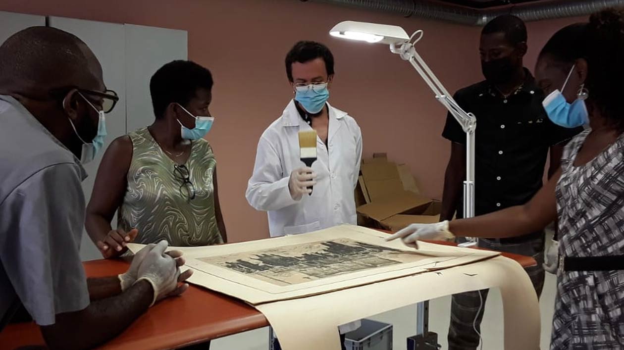Five people stand around a flat object in a restoration workshop, one person holds a brush