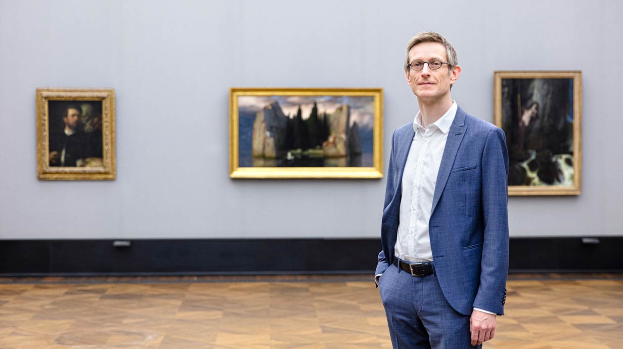 A man stands in an exhibition hall in front of three 19th century paintings