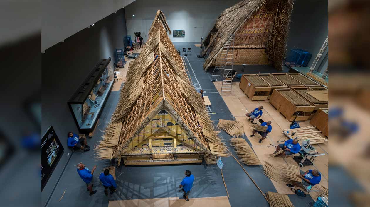 Dachdeckung im Humboldt Forum Mehrere Männer in blauen T-Shirts sind um ein traditionelles Haus aus Palau gruppiert, das sie mit Dachschindeln aus Palmblättern decken