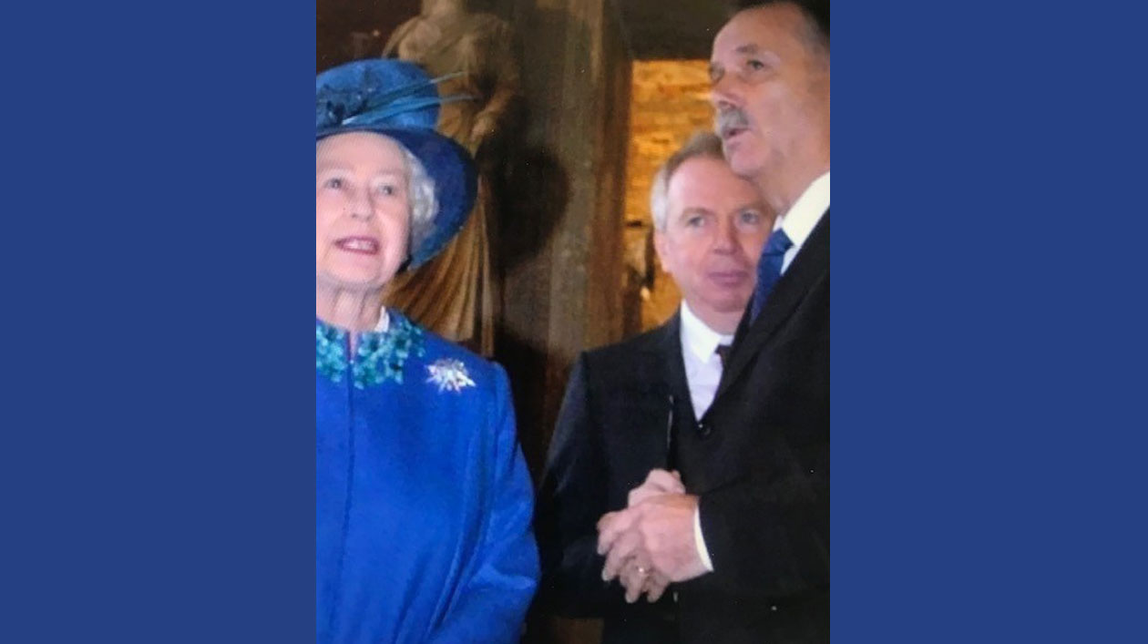 Three people stand side by side, one of them a queen, to her right two men in suits