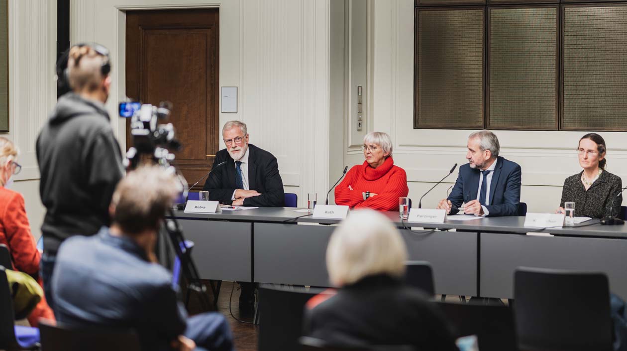 Fünf Personen sitzen an einem langen Tisch, vor ihnen sitzend Journalisten und Kamerateams