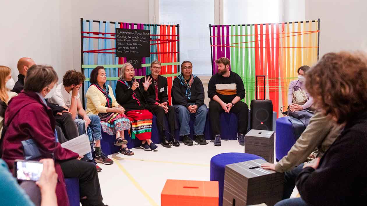 Several people sit in a semicircle in front of colorful ribbons and discuss, a woman speaks into a microphone