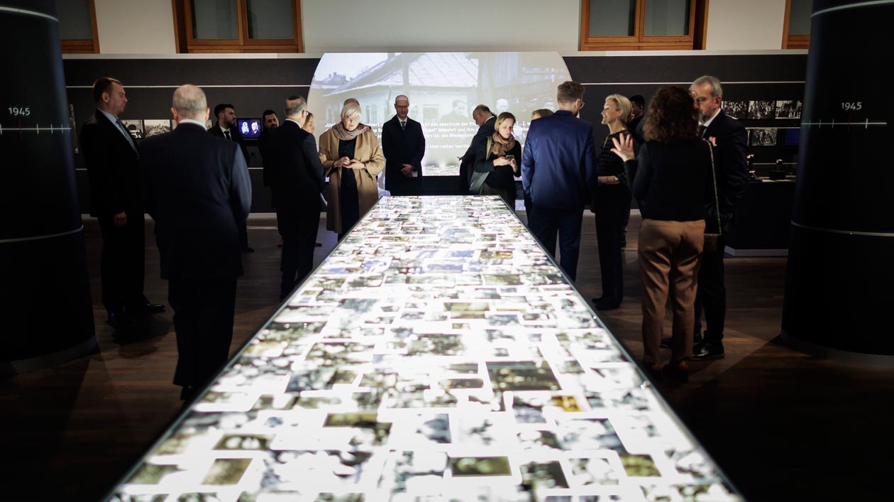 Auf einem Leuchttisch im Vordergrund liegen Fotografien, im Hintergrund unterhalten sich Menschen in einem Ausstellungsraum