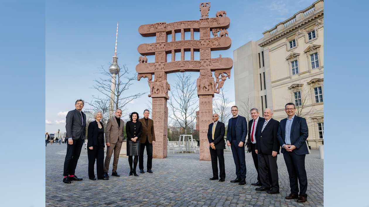 Group photo of 10 people in front of a gate of red sandstone; in the background is the Berlin TV Tower and the facade of the Humboldt Forum.