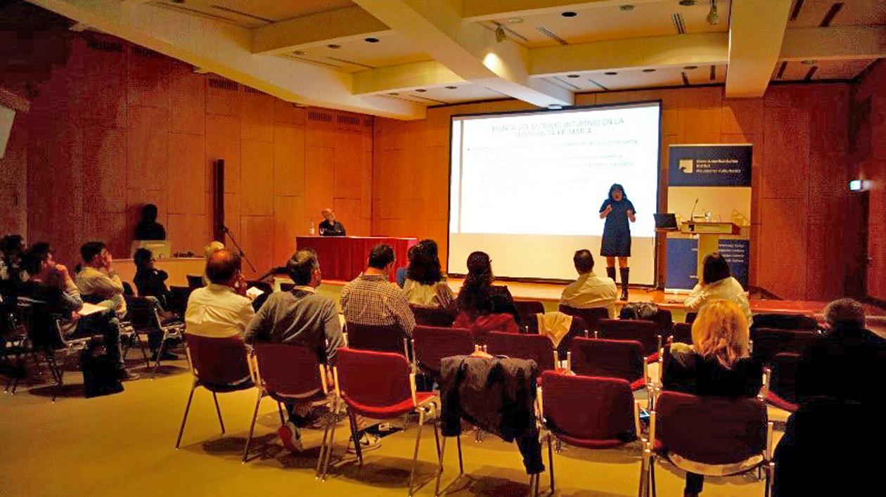 Konferenzraum mit Bühne und Leinwand, eine Person spricht zu anderen, die auf Stühlen sitzen