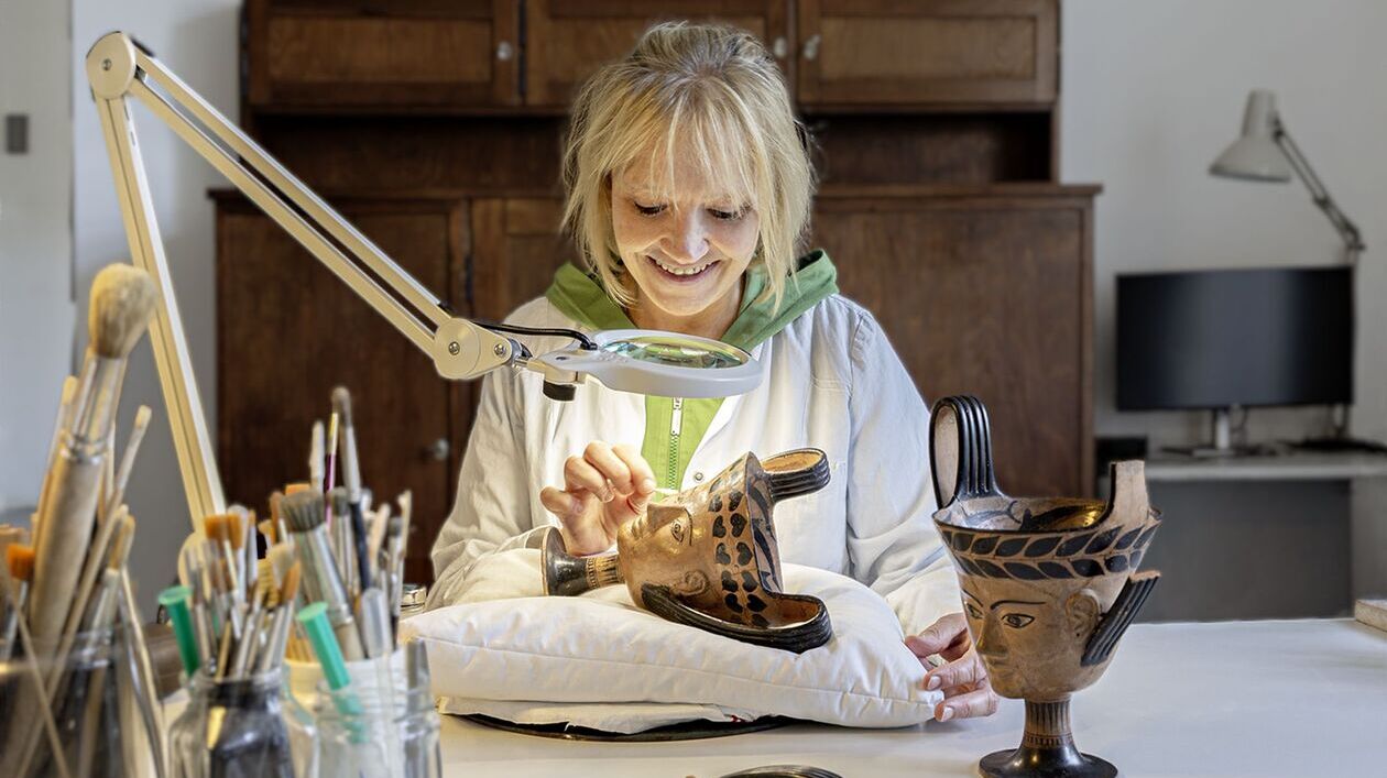 Under a magnifying lamp, Anke Kobbe is working on an antique ceramic vessel shaped like a face.