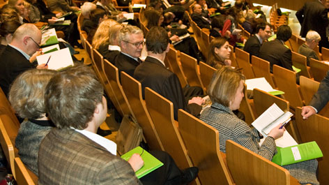 In Stuhlreihen eines Hörsaals sitzende Menschen mit grünen Veranstaltungsmappen