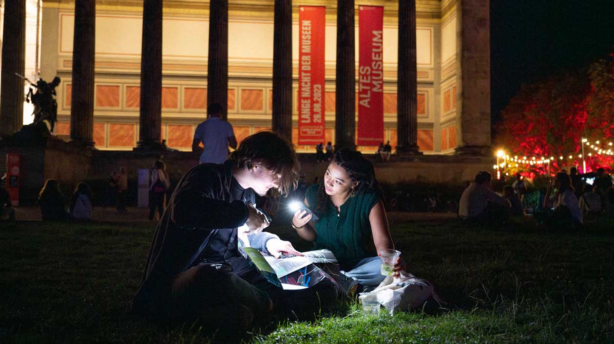 LNDM 2024, Lustgarten und Altes Museum Zwei junge Personen sitzen an Abend vor einem prachtvollen Museum und beleuchten mit Taschenlampen ein Programmheft